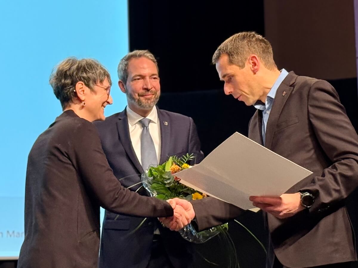 Zwei Männer und eine Frau, einer schüttelt der Frau die Hand und hält eine Urkunde in der Hand.