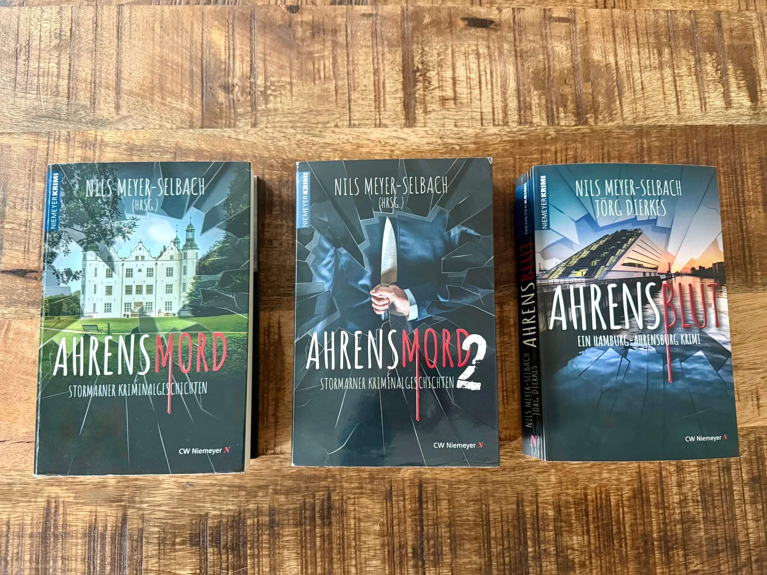 Drei Bücher auf einem Untergrund aus Holz: Ahrensmord, Ahrensmord 2, Ahrensblut