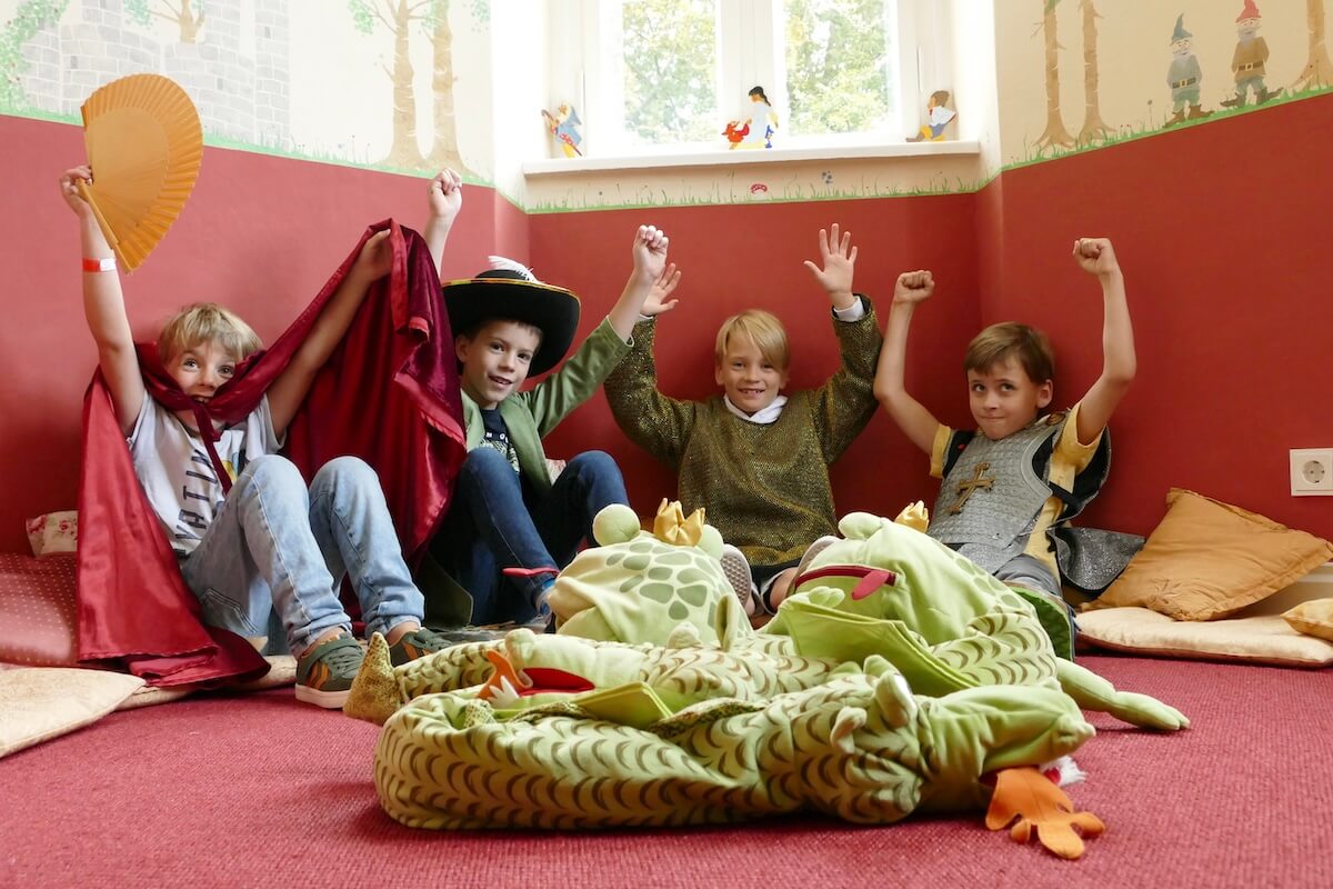 4 verkleidete Kinder sitzen auf dem Boden in einem Turmzimmer und heben die Hände über den Kopf