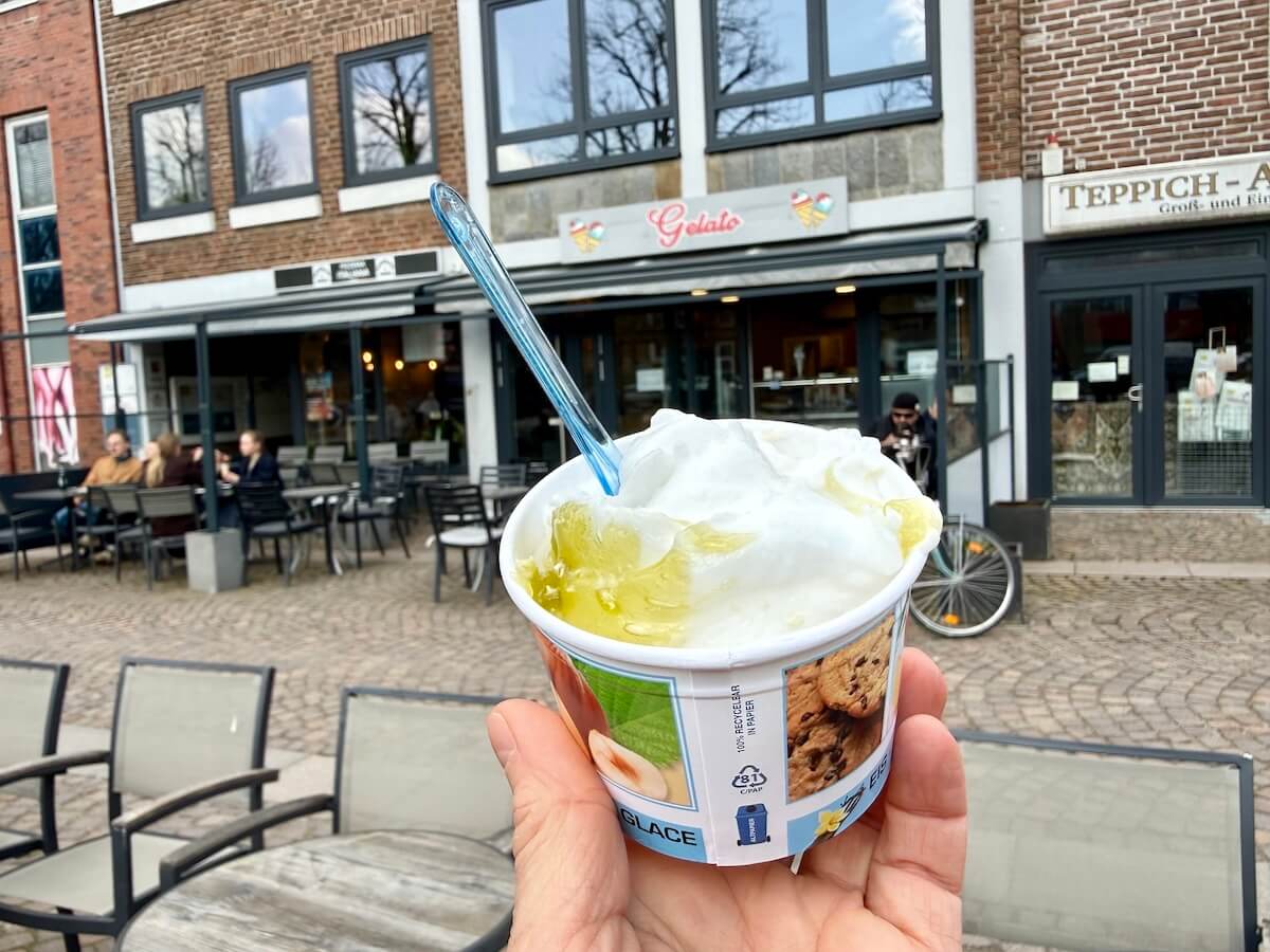 Ein Becher mit einer Kugel hellem Eis. Im Hintergrund ein Eiscafe