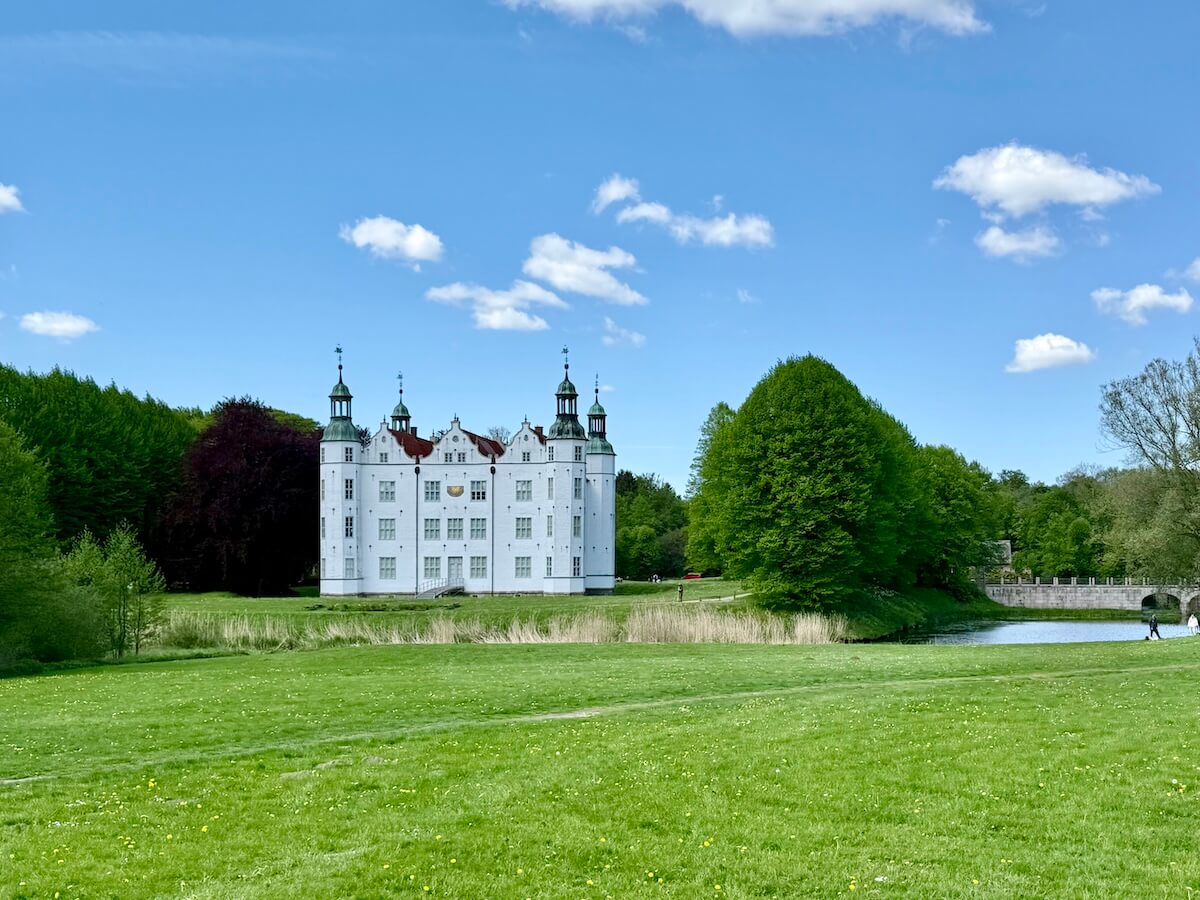 Weißes Schloss im Sommer, umgeben von Bäumen und Wiesen, blauer Himmel