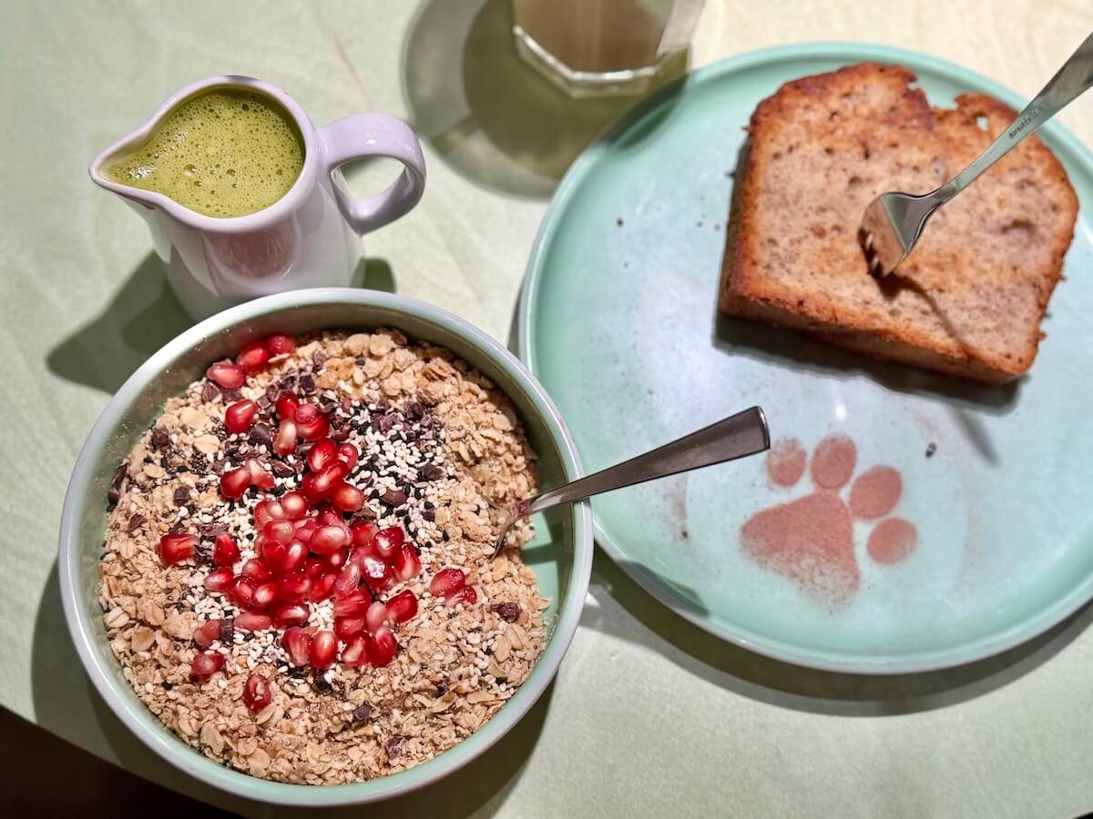 Eine Schale mit Müsli und ein Teller mit Bananenbrot von oben fotografiert. Auf dem Teller eine Pfote aus Kakaopulver